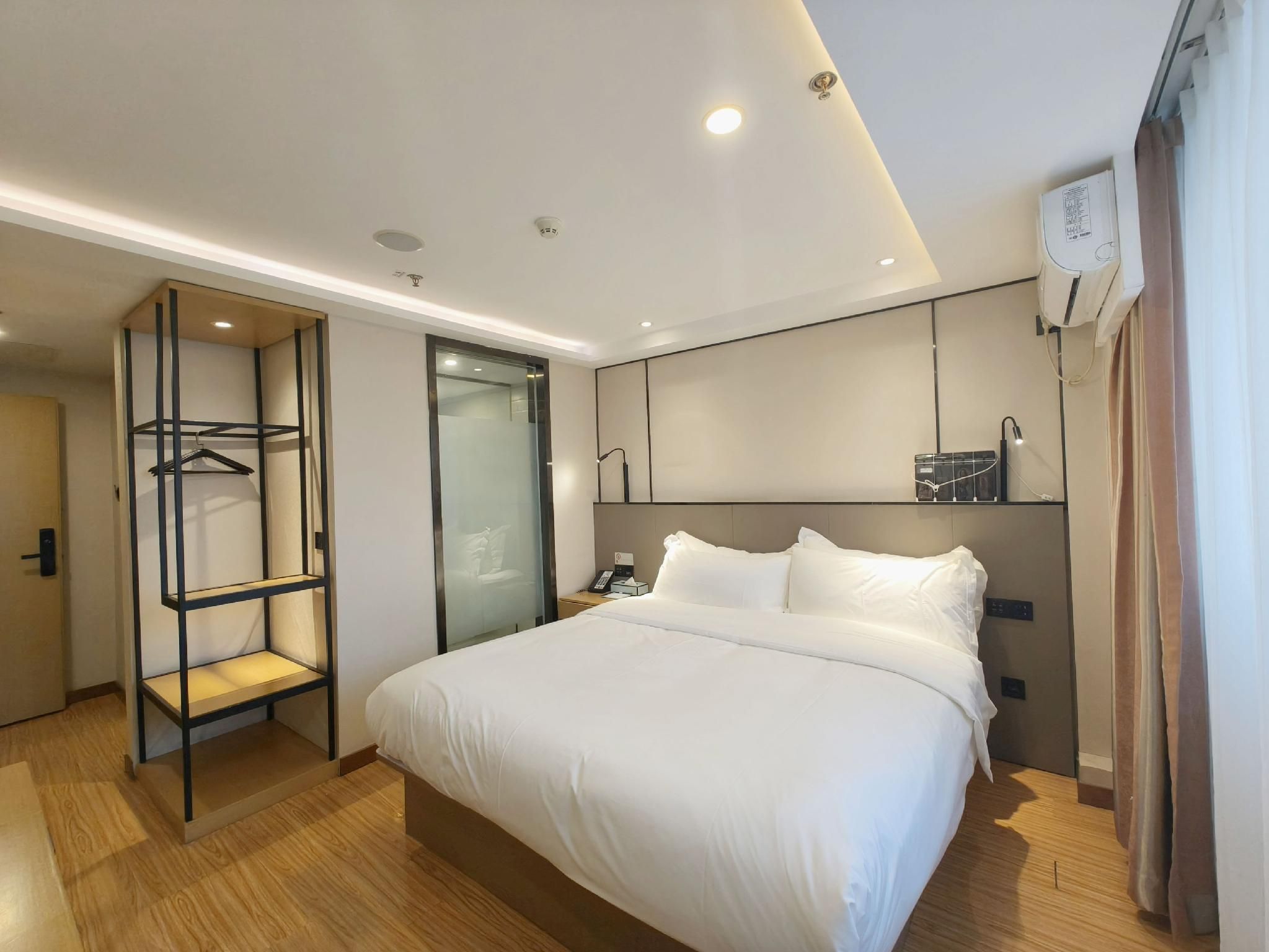 A modern hotel room with a large bed, a built-in wardrobe, a bedside table, and a wall-mounted air conditioning unit.