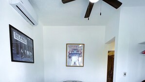 Een slaapkamer met een bed, een plafondventilator, een airconditioner, een ingelijst poster en een stoel.