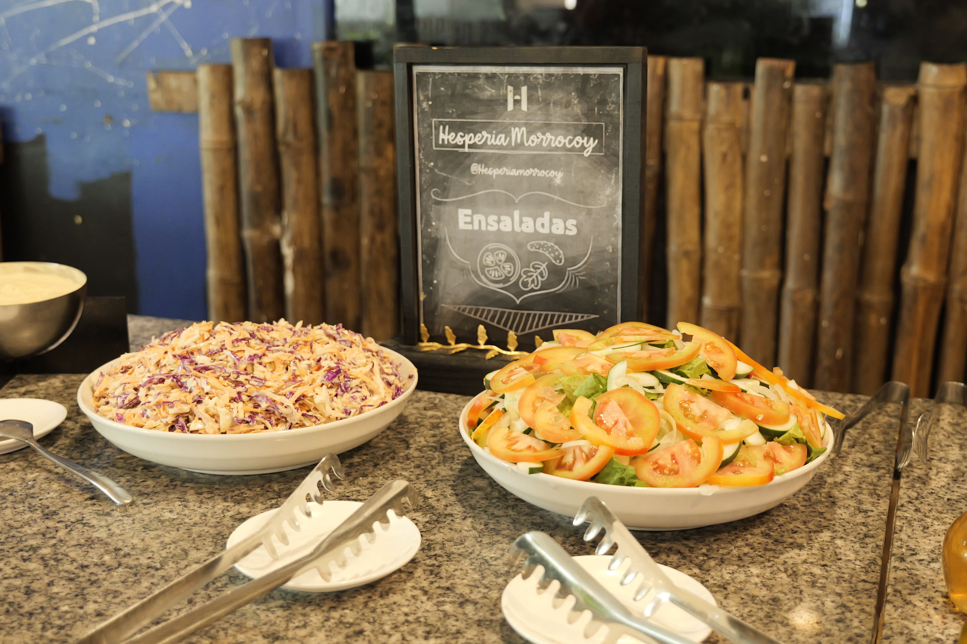 Bancada de restaurante com duas saladas em tigelas, um menu em lousa e talheres.