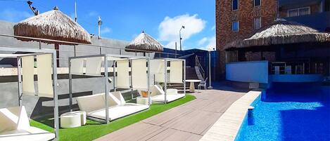 A swimming pool area with sun loungers and thatched umbrellas, adjacent to a high-rise building.