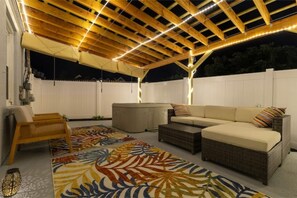 An outdoor patio area with a wooden pergola, cushioned seating, and a patterned rug.