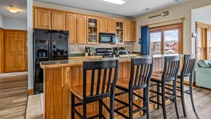 A kitchen with wooden cabinets, a black refrigerator, a microwave, and a bar area with chairs. - OBX Top-Floor Condo- Marina & Sound Views, Quiet & Serene (Manteo)