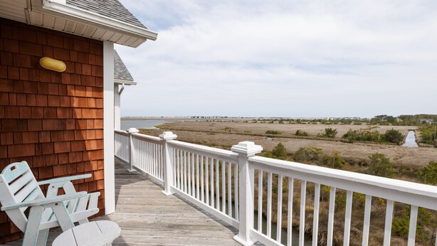A wooden deck with white railings, a table, and two chairs, overlooking a coastal landscape with a body of water and grassy fields. - OBX Top-Floor Condo- Marina & Sound Views, Quiet & Serene (Manteo)