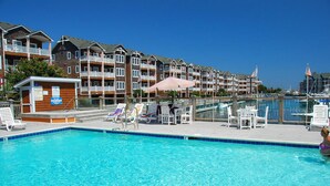 A swimming pool with lounge chairs and umbrellas, surrounded by multi-story residential buildings with balconies. - OBX Top-Floor Condo- Marina & Sound Views, Quiet & Serene (Manteo)