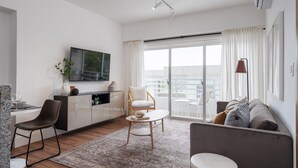 A modern living room with a sofa, a coffee table, a dining area with a chair, a TV mounted on the wall, and a large sliding glass door leading to a balcony.