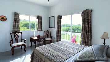 A bedroom with a bed, two chairs, a small table, a ceiling fan, and large windows with curtains.