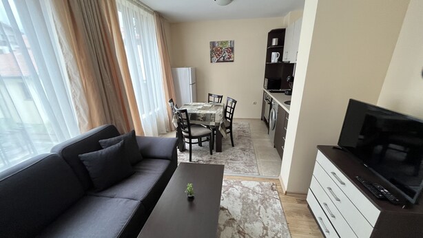 A modern living room with a sofa, a coffee table, and a TV stand. There is a dining area with a table and chairs. The room has a kitchen area with a microwave and a refrigerator.