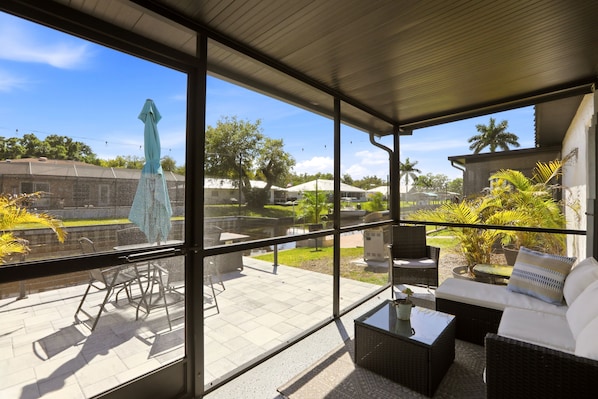 A covered patio with outdoor furniture, a glass wall, and a view of a landscaped garden.