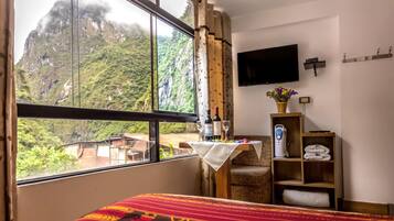 Una habitación con una cama, un televisor, una mesa con una botella y vasos, un estante con toallas y vistas a una montaña.