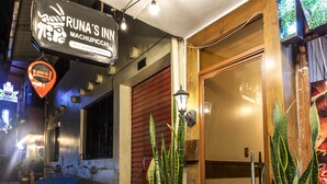 Entrada de hotel com uma placa indicando "Runa's Inn" e serviço de "Bed & Breakfast". Há plantas em vasos e um carrinho azul na parte externa.