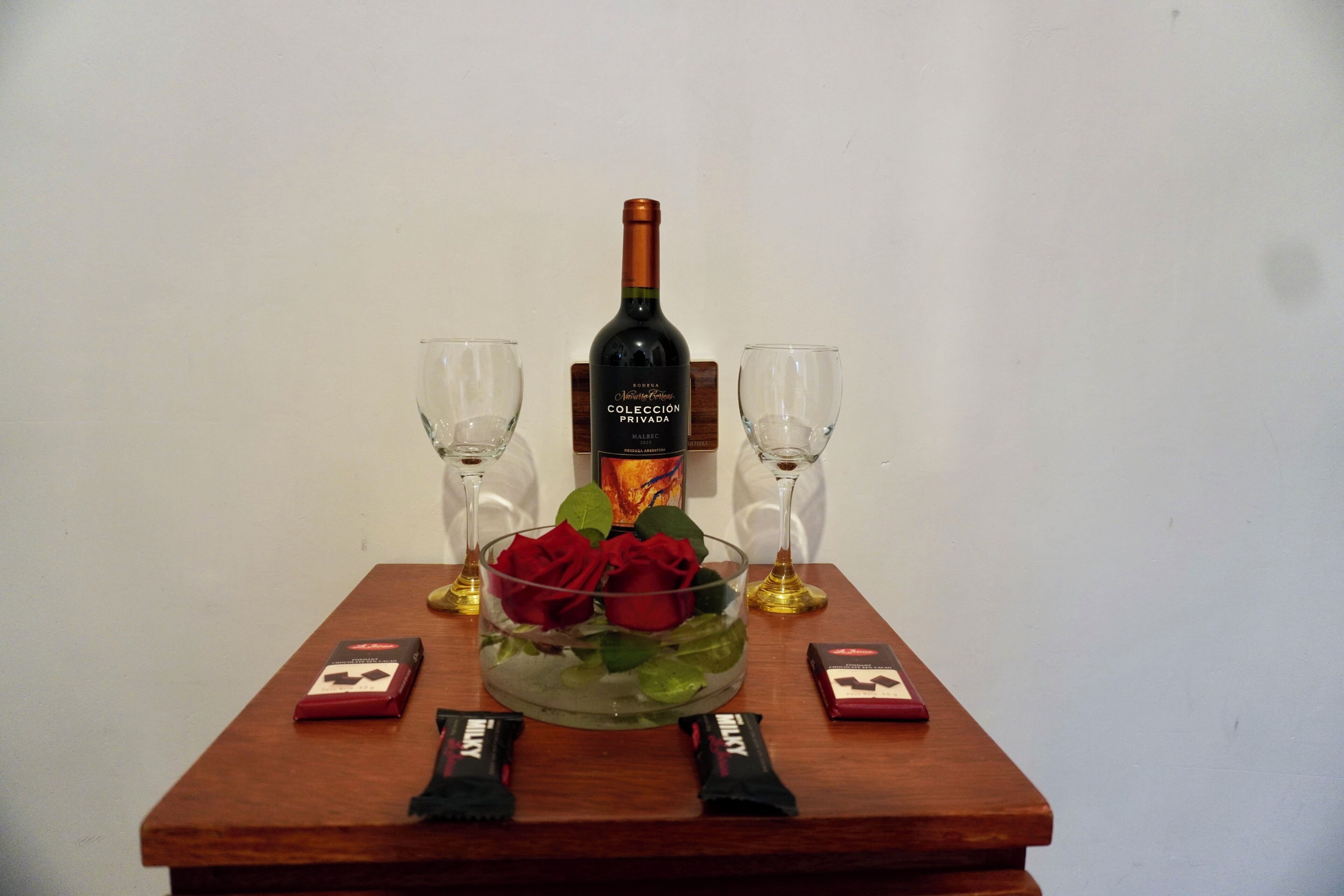 A wooden table with a bottle of wine, two glasses, a bowl with roses, and two chocolate bars.