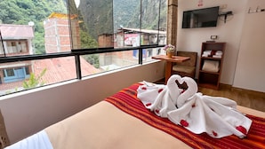 Quarto de hotel com uma grande janela com vista para uma paisagem montanhosa. Há uma cama com colcha de padrão vermelho e bege e uma toalha branca disposta em forma de coração. Uma pequena mesa de madeira com um vaso de flores está colocada ao lado da cama.
