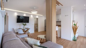 A modern living room with a sofa, dining table, and chairs. There is a TV mounted on the wall and a kitchen area visible through an open door. - Villa Mediluzma Almuñécar by Ruralidays (Granada)