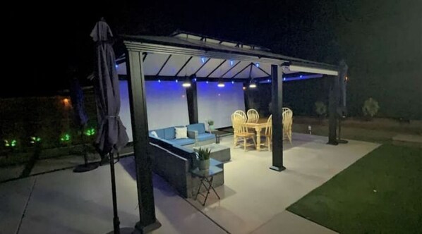 Un patio exterior con una zona de estar techada, una mesa y sillas.
