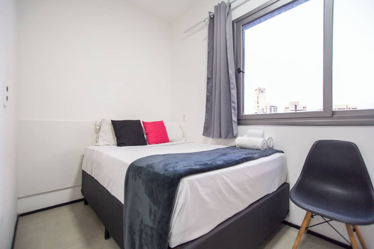 Quarto de hotel com cama, cadeira, janela com cortinas e vista para prédios.