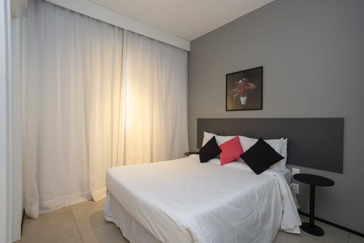 Cama bem arrumada com roupa de cama branca e almofadas pretas e vermelhas, uma mesa de cabeceira e um quadro na parede.