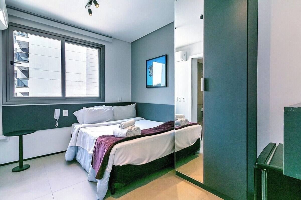Quarto de hotel moderno com cama, mesa de cabeceira, janela e espelho.