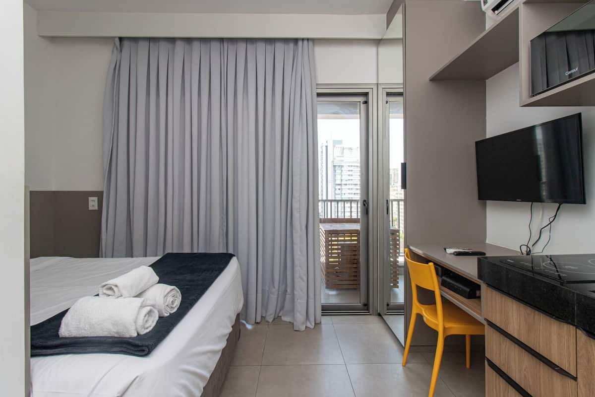 Quarto de hotel com cama, escrivaninha, cadeira, televisão e vista da cidade pela janela.