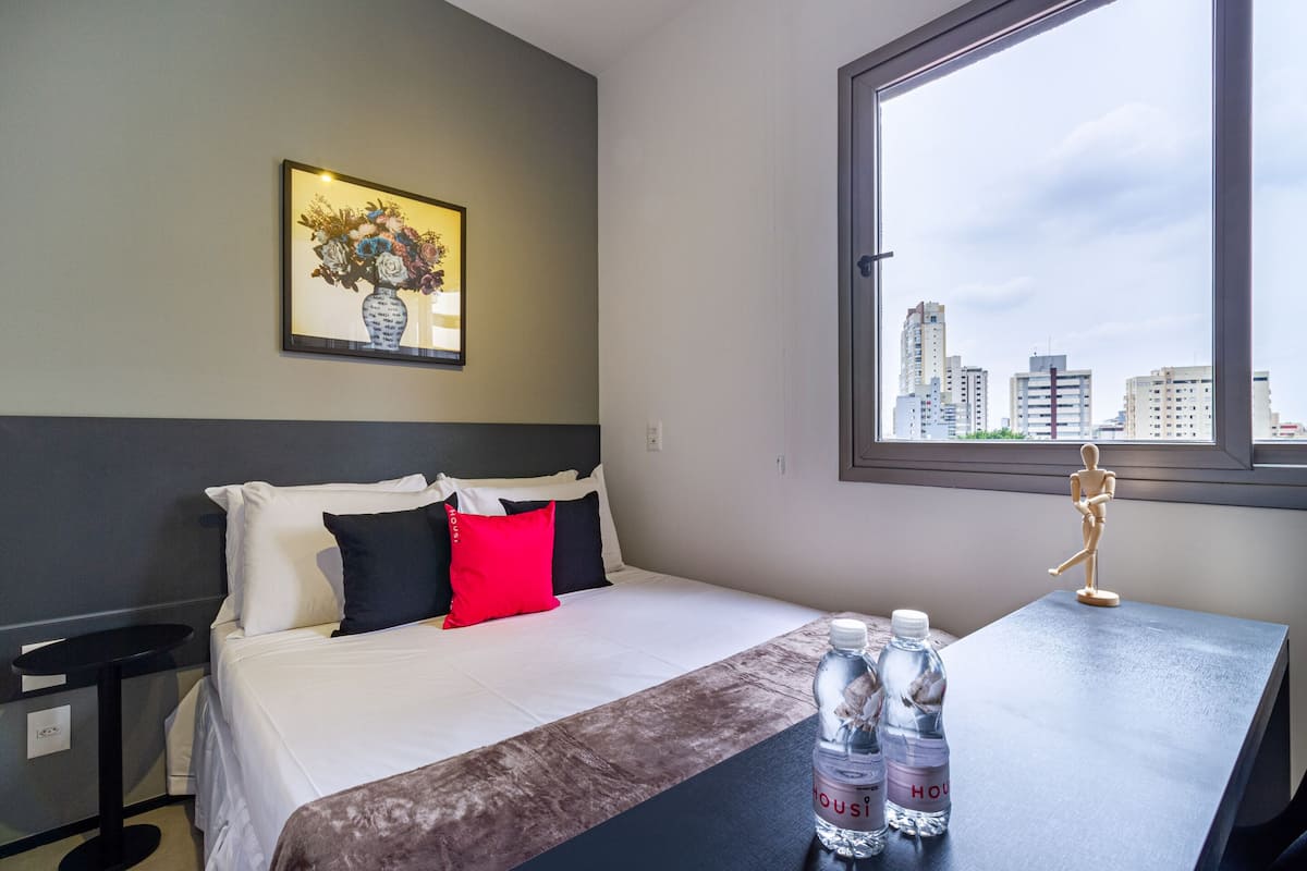 Quarto de hotel com uma cama, uma pequena mesa com garrafas de água, uma janela com vista para a cidade e um quadro na parede.
