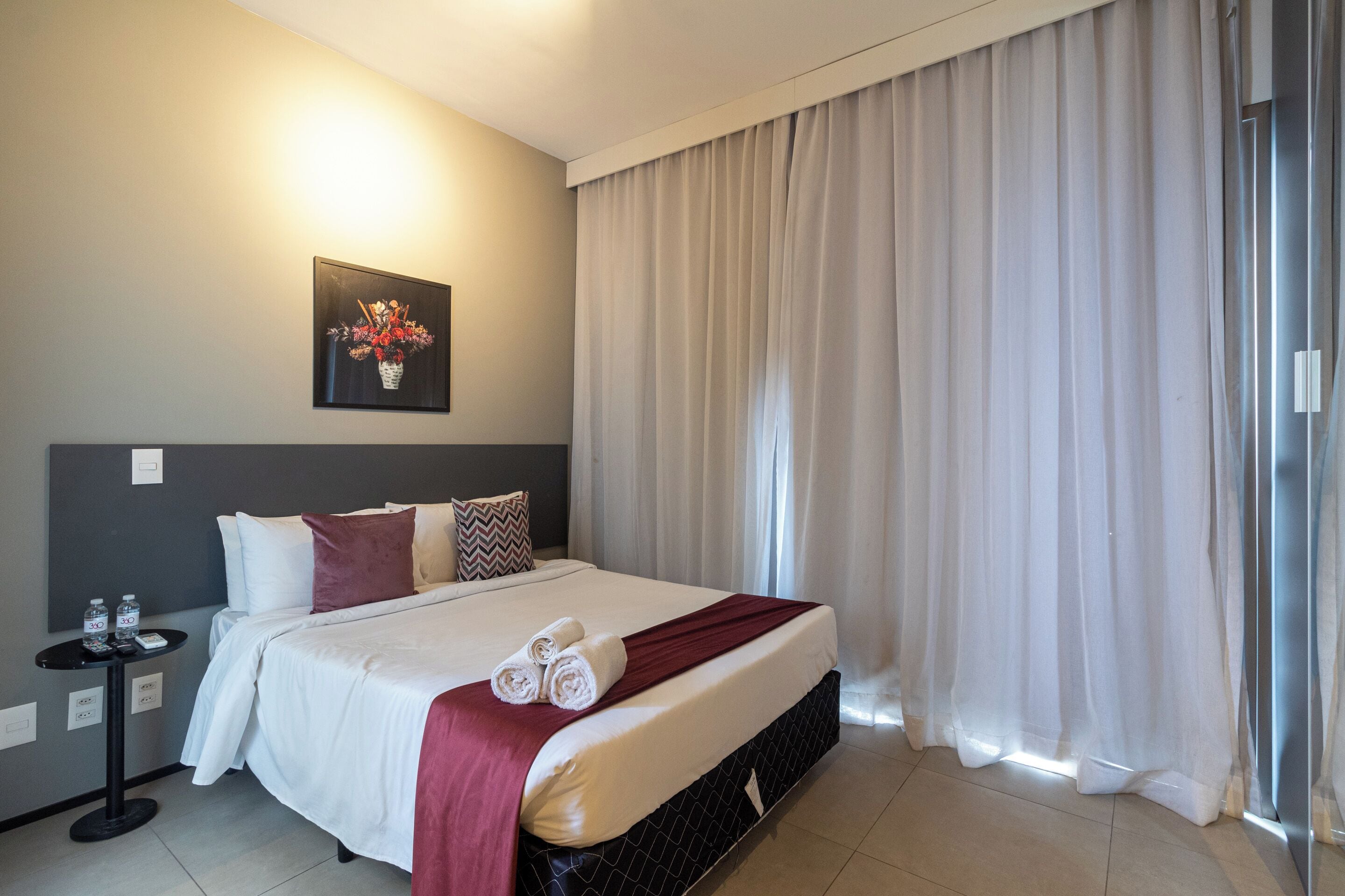 A hotel room with a neatly made bed, a small table with a phone and water bottles, and a painting on the wall.
