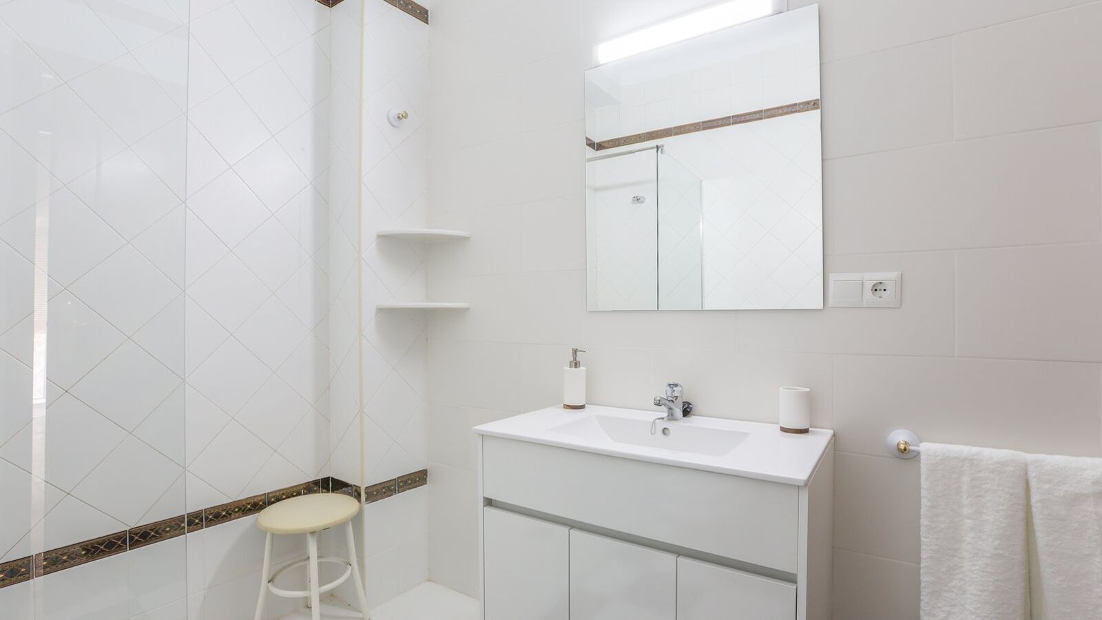 Un baño moderno con azulejos blancos, cabina de ducha de cristal, lavamanos blanco con espejo encima y un taburete blanco.