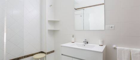 Un baño moderno con azulejos blancos, cabina de ducha de cristal, lavamanos blanco con espejo encima y un taburete blanco.