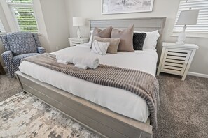Une chambre à coucher avec un grand lit, un fauteuil à motifs, deux tables de chevet avec des lampes et un tableau encadré au mur.