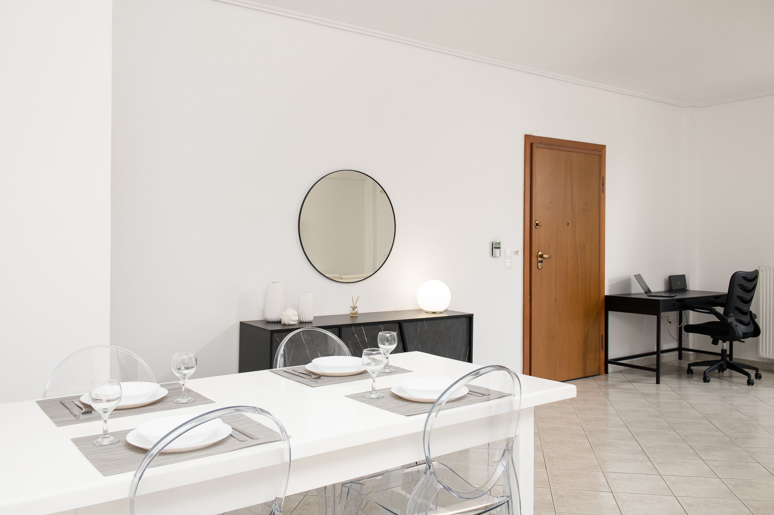 A modern dining area with a white table set for two, clear chairs, a black console table with a mirror, and a wooden door in the background.