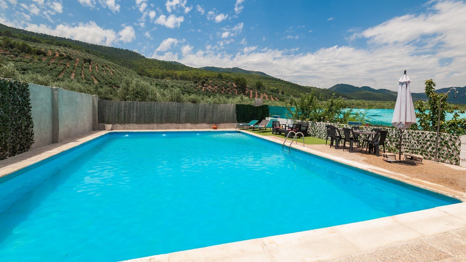 A rectangular swimming pool with a clear blue water surface, surrounded by a stone deck. There are lounge chairs and umbrellas on the right side, and a view of a hilly landscape in the background.
