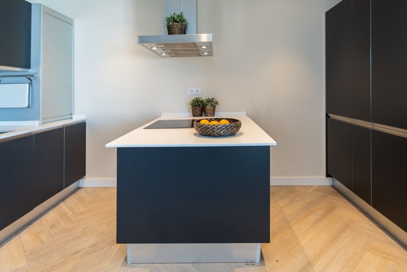 A modern kitchen with dark cabinetry, a white countertop, and a central island with a bowl of fruit.