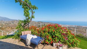 A stone bench with a cushion and a book, surrounded by vibrant red and pink flowers, overlooking a coastal landscape.
