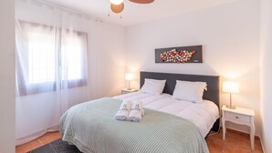 A neatly made bed with white linens and a green striped blanket, two bedside tables with lamps, a window with sheer curtains, and a wall-mounted artwork.
