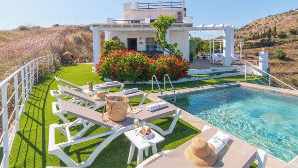 A poolside area with lounge chairs, a small table with a drink and a hat, and a building with a garden in the background.