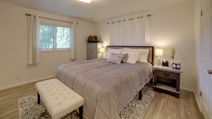 A bedroom with a large bed, a bench, a nightstand, and a window with curtains. - Simple + Charming Idaho Retreat w/ Patio! (Idaho Falls)