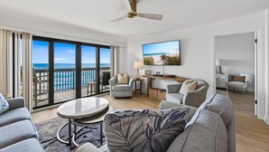 A spacious living room with a balcony overlooking the ocean, a ceiling fan, and a flat-screen TV mounted on the wall.