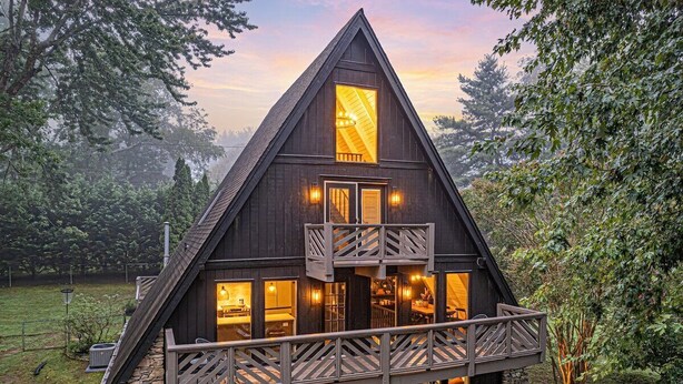 A dark wooden cabin with a triangular roof, a balcony with railings, and a lit interior visible through the windows. - Modern A-Frame, Hot Tub, EV, 10 Guests, Dual King, 8 Mins from Downtown Hendersonville (Hendersonville)