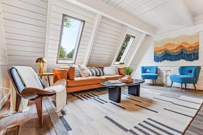 A cozy loft-style room with a sloped ceiling, a sofa, two armchairs, a coffee table, and a large window.