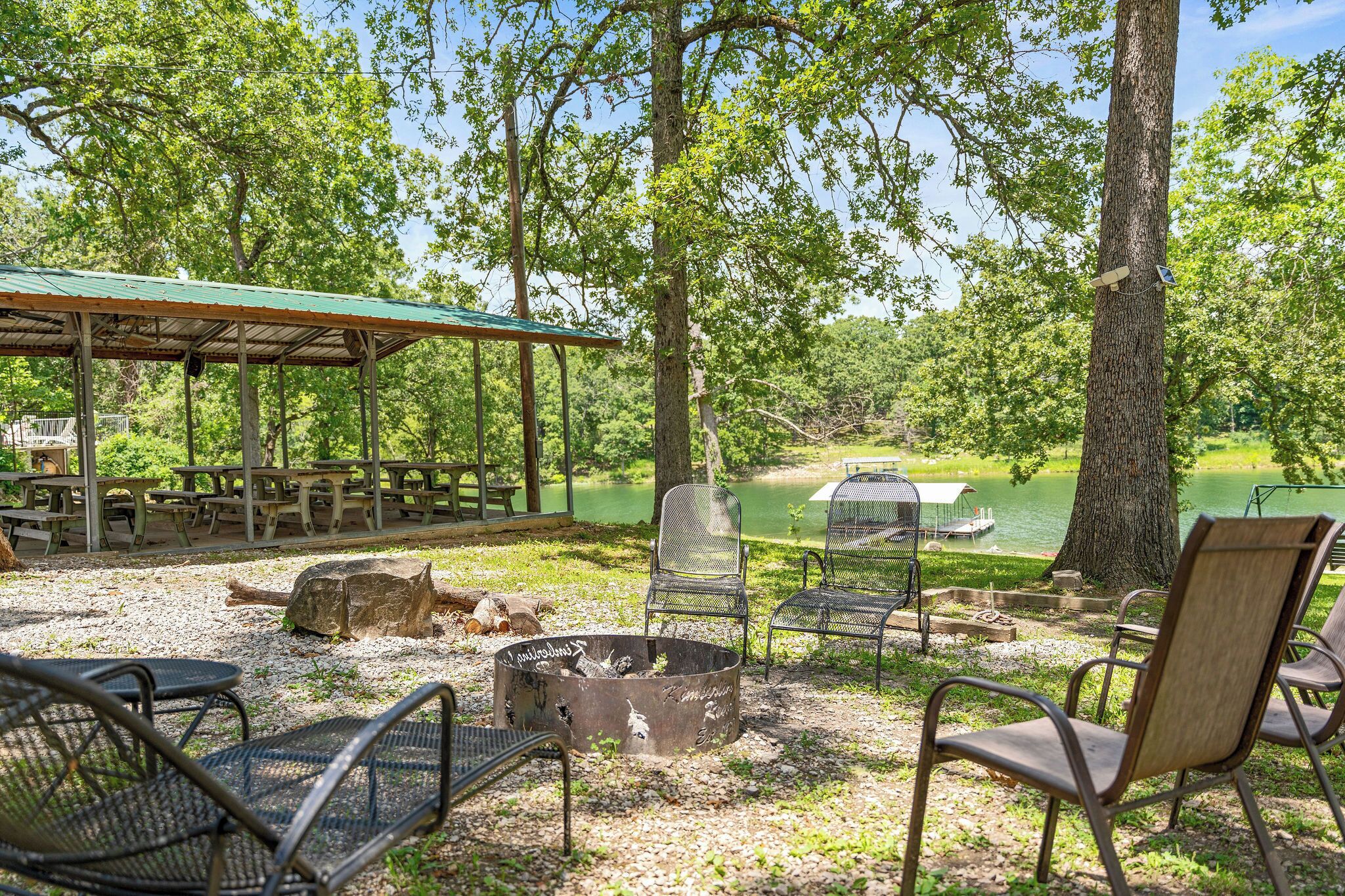 A covered picnic area with metal chairs, a fire pit, and a view of a lake.