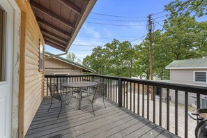 Terraza de madera con mesa y sillas de metal, un espacio cubierto y vistas a árboles y otro edificio.