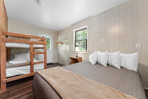 Una habitación con una cama individual doble, una cama individual con almohadas blancas, una mesita de noche de madera, una lámpara y una ventana con persianas.
