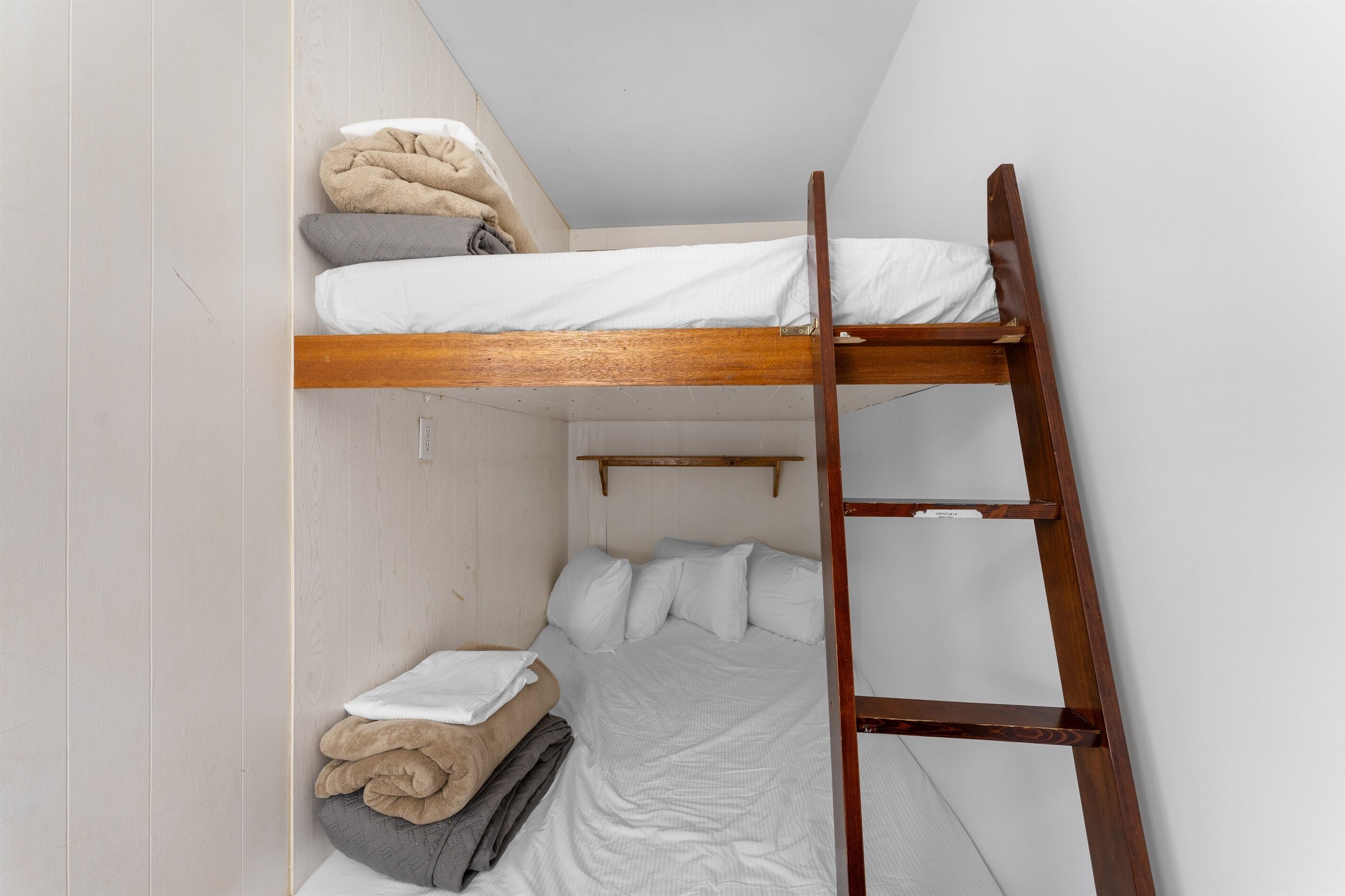 Una cama litera con escalera de madera, ropa de cama blanca y un estante pequeño sobre la cama inferior.