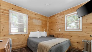 Quarto com painéis de madeira, uma cama, televisão fixada na parede e janela com persianas.