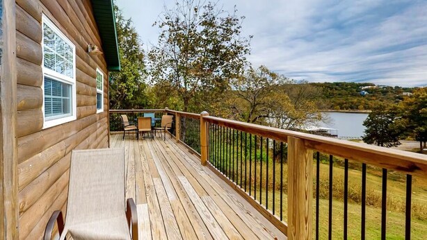 A wooden deck with a railing, a table, and chairs, overlooking a lake and trees. - 2BR Waterfront Duplex Cabin w/ Pool + Swim & Boat dock (Branson)