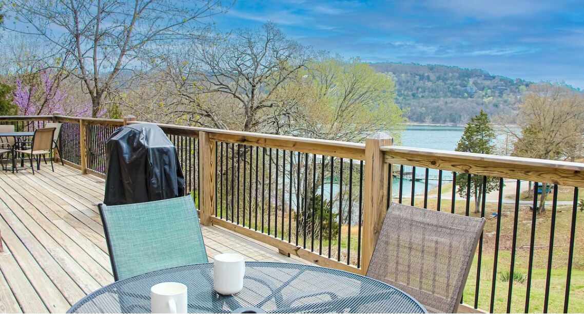 Una terraza de madera con mobiliario exterior, con vistas a un lago y colinas.