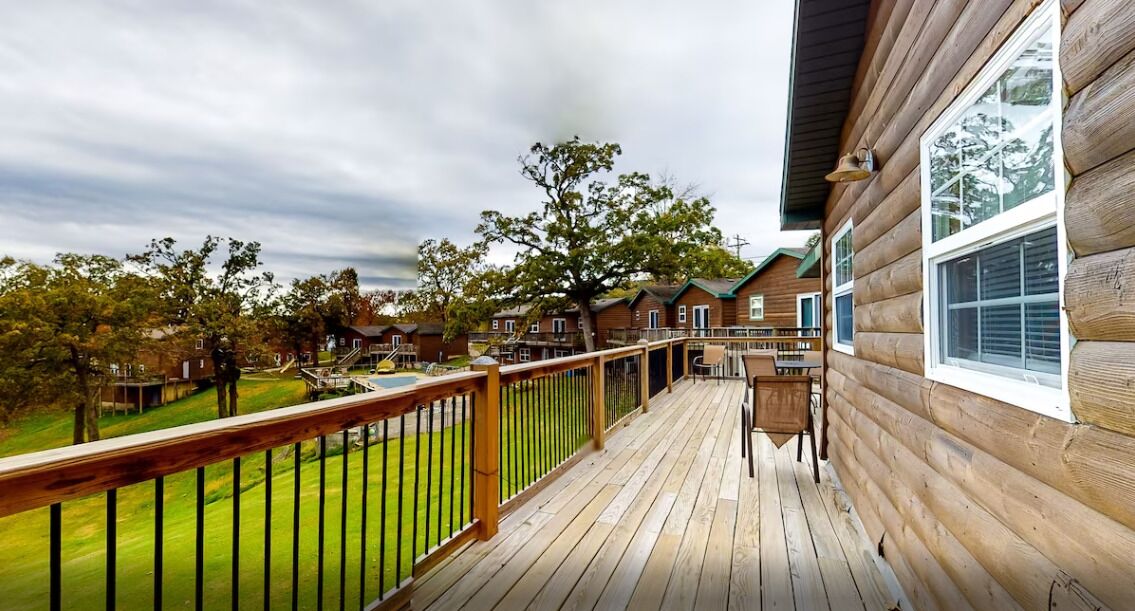 Une terrasse en bois, dotée d’une rambarde, qui donne sur un espace verdoyant agrémenté d’arbres et d’autres bâtiments.