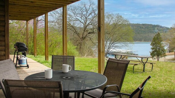 Une terrasse couverte avec une table et des chaises, donnant sur un lac et une aire de pique-nique.