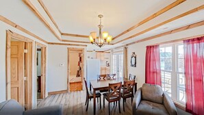 Un coin repas avec une table et des chaises en bois, un lustre et une vue sur la cuisine par une porte ouverte.