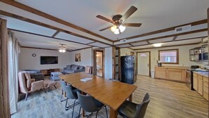 Un vaste espace de vie comprenant une table à manger en bois, des sièges confortables, un ventilateur de plafond et un coin cuisine doté de meubles en bois.