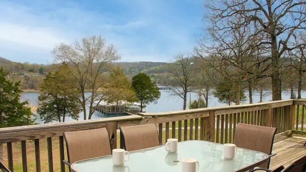 Une terrasse dotée d’une table et de chaises en verre, offrant une vue sur un lac et des arbres.