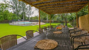 Un espace extérieur aménagé pour les repas, avec une pergola, des chaises en osier, une table dressée avec des sets de table et des fruits, et une piscine en arrière-plan.